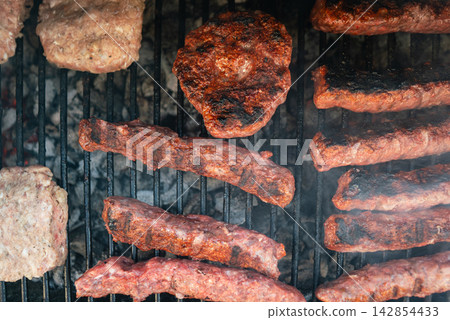 Meat patties and sausage rolls cooking on grill over charcoal. Smoky BBQ scene 142854433