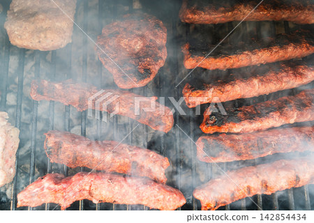Meat patties and sausage rolls cooking on grill over charcoal. Smoky BBQ scene Meat patties and sausage rolls cooking on grill over charcoal. Smoky BBQ scene 142854434