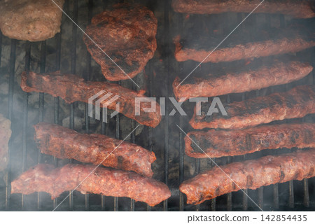 Meat patties and sausage rolls cooking on grill over charcoal. Smoky BBQ scene 142854435