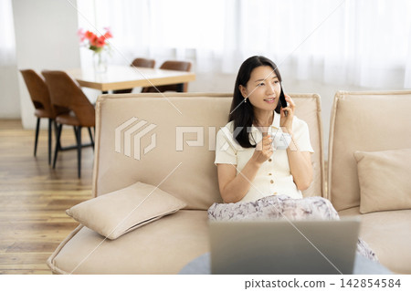一位年輕的日本女子坐在窗邊的沙發上,手裡拿著咖啡杯,正在用智慧型手機通話。 一位年輕的日本女子坐在窗邊的沙發上,手裡拿著咖啡杯,正在用智慧型手機通話。 142854584
