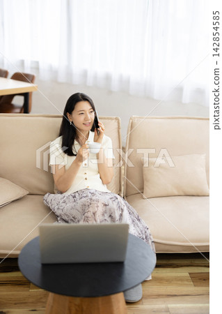 A young Japanese woman is sitting on a sofa by the window, holding a coffee cup and talking on her smartphone. A young Japanese woman is sitting on a sofa by the window, holding a coffee cup and talking on her smartphone. 142854585