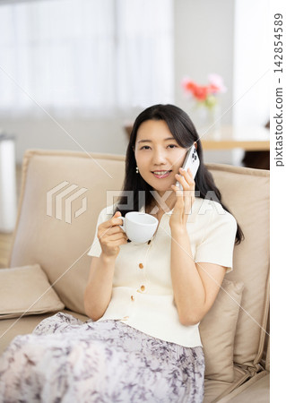 A young Japanese woman is sitting on a sofa by the window, holding a coffee cup and talking on her smartphone. A young Japanese woman is sitting on a sofa by the window, holding a coffee cup and talking on her smartphone. 142854589
