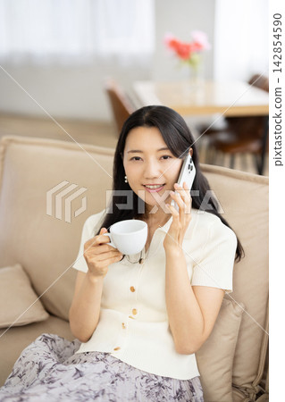 A young Japanese woman is sitting on a sofa by the window, holding a coffee cup and talking on her smartphone. A young Japanese woman is sitting on a sofa by the window, holding a coffee cup and talking on her smartphone. 142854590