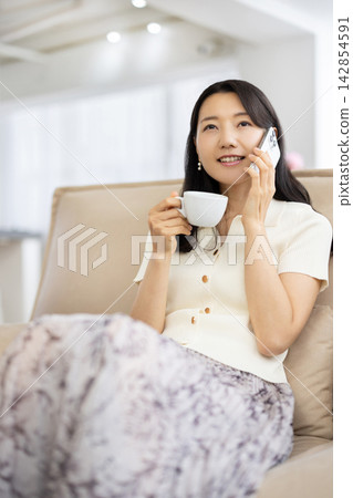 A young Japanese woman is sitting on a sofa by the window, holding a coffee cup and talking on her smartphone. A young Japanese woman is sitting on a sofa by the window, holding a coffee cup and talking on her smartphone. 142854591