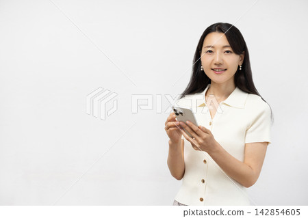 一位年輕的日本女子手持智慧型手機，站在白牆前對著鏡頭微笑。 142854605