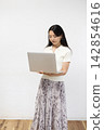 A young Japanese woman is using a laptop in front of a white wall. 142854616