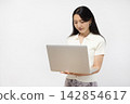 A young Japanese woman is using a laptop in front of a white wall. 142854617