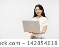 A young Japanese woman smiles at the camera, holding a laptop, in front of a white wall. 142854618