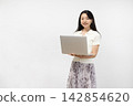 A young Japanese woman smiles at the camera, holding a laptop, in front of a white wall. 142854620