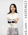 A young Japanese woman smiles at the camera, holding a gift box in front of a white wall. 142854623