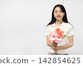 A young Japanese woman smiles at the camera, holding a bouquet of flowers, in front of a white wall. 142854625