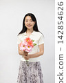 A young Japanese woman smiles at the camera, holding a bouquet of flowers, in front of a white wall. 142854626