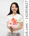 A young Japanese woman smiles at the camera, holding a bouquet of flowers, in front of a white wall. 142854627