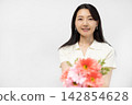 A young Japanese woman smiles at the camera, holding a bouquet of flowers, in front of a white wall. 142854628