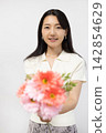 A young Japanese woman smiles at the camera, holding a bouquet of flowers, in front of a white wall. 142854629
