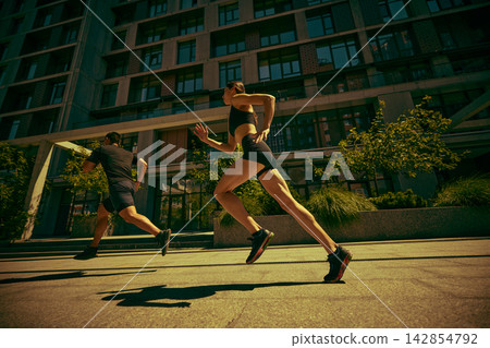 Young athletic pair races past modern buildings with determined motivation Young athletic pair races past modern buildings with determined motivation 142854792