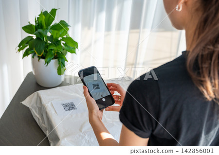 Person scanning QR code on parcel with smartphone camera for delivery tracking at home. Contactless technology mobile app and digital service concept. Person scanning QR code on parcel with smartphone camera for delivery tracking at home. Contactless technology mobile app and digital service concept. 142856001