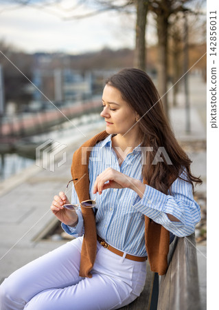 Elegant young woman in old money style sitting on bench near pier in Prague holding sunglasses in early spring. Understated luxury and timeless European femininity concept. Elegant young woman in old money style sitting on bench near pier in Prague holding sunglasses in early spring. Understated luxury and timeless European femininity concept. 142856011