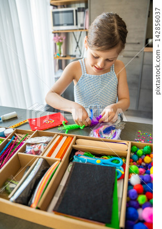 Little girl gluing colorful feathers to handmade craft toy at table with creative supplies. Kids creativity fine motor skills development and learning through play concept. Little girl gluing colorful feathers to handmade craft toy at table with creative supplies. Kids creativity fine motor skills development and learning through play concept. 142856037
