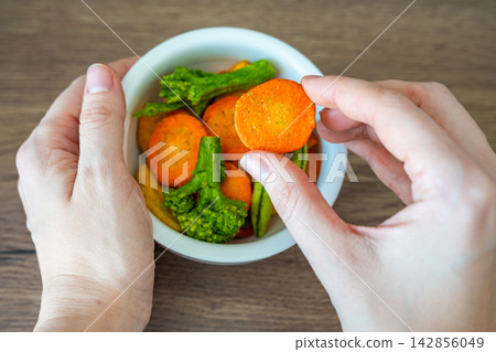 Hand taking carrot vegetable chip from bowl on wooden table. Healthy snack choice and clean eating lifestyle concept. Hand taking carrot vegetable chip from bowl on wooden table. Healthy snack choice and clean eating lifestyle concept. 142856049