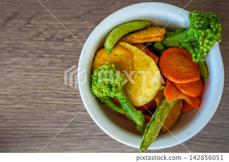 Mixed vegetable chips in bowl with broccoli, carrot, peas and sweet potato on wooden table. Healthy snack and clean eating concept without salt sugar or additives. Mixed vegetable chips in bowl with broccoli, carrot, peas and sweet potato on wooden table. Healthy snack and clean eating concept without salt sugar or additives. 142856051