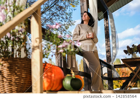 Woman in cozy sweater drinking warm tea on wooden cottage porch with autumn decorations and pumpkins, enjoying sunny morning weather Woman in cozy sweater drinking warm tea on wooden cottage porch with autumn decorations and pumpkins, enjoying sunny morning weather 142858564
