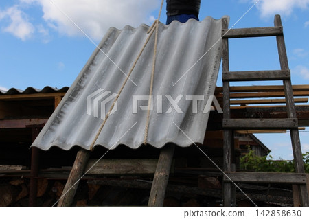 Worker pulling a corrugated slate sheet up a wooden ladder using a rope against a blue sky Worker pulling a corrugated slate sheet up a wooden ladder using a rope against a blue sky 142858630