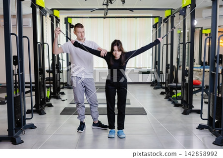 Physiotherapist assisting young woman exercising with cable machine in rehabilitation center Physiotherapist assisting young woman exercising with cable machine in rehabilitation center 142858992