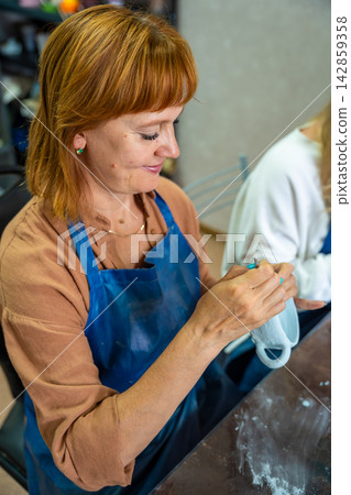 Woman applying blue glaze to handmade ceramic bowl with brush in pottery workshop. Ceramic craft process represents artisan work and creative hobby Woman applying blue glaze to handmade ceramic bowl with brush in pottery workshop. Ceramic craft process represents artisan work and creative hobby 142859358