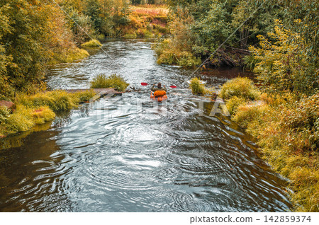 Individual navigating small bright orange boat through turbulent dark creek passing ruined stone structure within dense yellowing foliage during daytime Individual navigating small bright orange boat through turbulent dark creek passing ruined stone structure within dense yellowing foliage during daytime 142859374