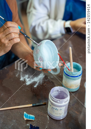 Woman painting floral details on handmade ceramic mug with fine brush. Detailed pottery decoration represents craftsmanship and creative ceramic art Woman painting floral details on handmade ceramic mug with fine brush. Detailed pottery decoration represents craftsmanship and creative ceramic art 142859414