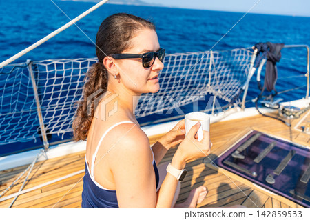 Woman enjoying a cup of coffee on the deck of a sailing yacht while cruising across the sea. Concept of everyday routine, life aboard, freedom, relaxation and long-term sailing travel. Woman enjoying a cup of coffee on the deck of a sailing yacht while cruising across the sea. Concept of everyday routine, life aboard, freedom, relaxation and long-term sailing travel. 142859533