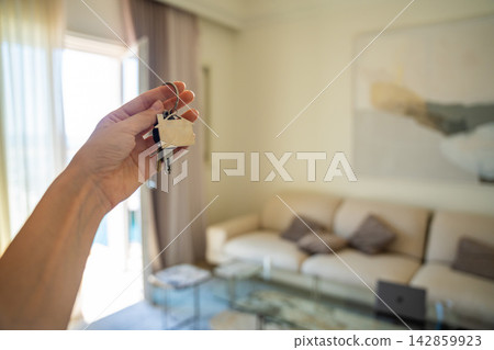 Hand holding a house key with a blurred modern apartment interior in the background. Concept of buying real estate, moving into a new home and property ownership. 142859923