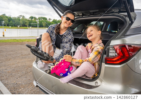 Mother and daughter putting on inline skates while sitting in the open car trunk before skating together. Active family lifestyle and quality time encourage healthy childhood and outdoor recreation Mother and daughter putting on inline skates while sitting in the open car trunk before skating together. Active family lifestyle and quality time encourage healthy childhood and outdoor recreation 142860127