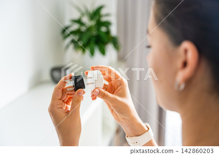 Woman compares electrical plug adapters for different power outlets at home. International travel and power compatibility concept for global electronics. Woman compares electrical plug adapters for different power outlets at home. International travel and power compatibility concept for global electronics. 142860203