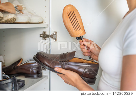 Person holds a leather shoe and a wooden shoe tree near an open wardrobe. Regular shoe care supports seasonal storage and helps retain footwear form. Person holds a leather shoe and a wooden shoe tree near an open wardrobe. Regular shoe care supports seasonal storage and helps retain footwear form. 142860529