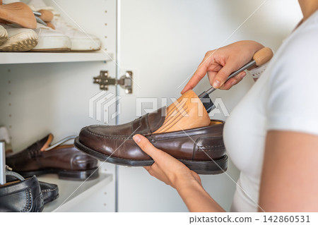 Person inserts a wooden shoe tree into a leather loafer beside a wardrobe. Crease prevention and shape retention improve long term footwear storage. 142860531