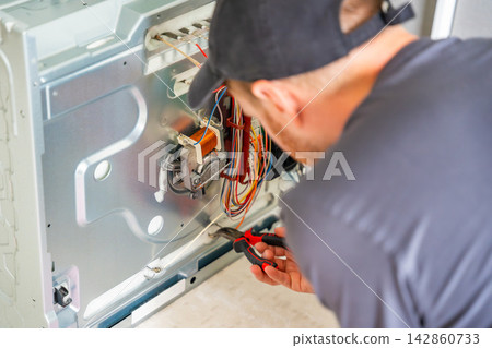 Technician servicing internal electrical components of disassembled electric oven during home appliance repair. Professional maintenance concept Technician servicing internal electrical components of disassembled electric oven during home appliance repair. Professional maintenance concept 142860733
