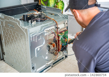 Technician testing electronic components and wiring inside disassembled electric oven. Appliance diagnostics and professional repair service concept Technician testing electronic components and wiring inside disassembled electric oven. Appliance diagnostics and professional repair service concept 142860734