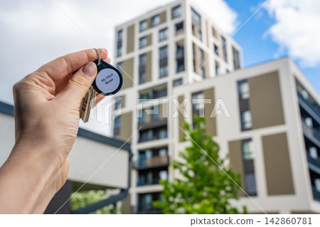 Hand holding house keys with My First Home keychain near modern apartment building. Property ownership and first real estate purchase concept Hand holding house keys with My First Home keychain near modern apartment building. Property ownership and first real estate purchase concept 142860781