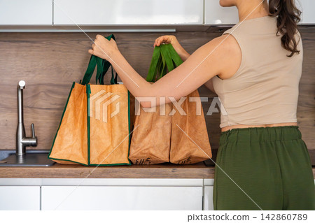 Woman carrying reusable grocery bags to kitchen after home delivery. Convenient food shopping and sustainable lifestyle concept Woman carrying reusable grocery bags to kitchen after home delivery. Convenient food shopping and sustainable lifestyle concept 142860789
