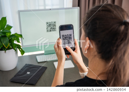 Person scanning QR code from computer screen with smartphone at home workspace. Digital interaction between devices and online service access concept. Person scanning QR code from computer screen with smartphone at home workspace. Digital interaction between devices and online service access concept. 142860934