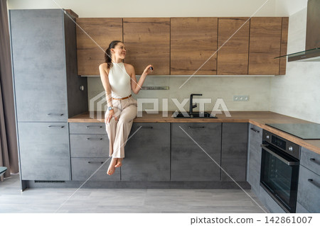 Young woman sitting on kitchen counter in modern apartment and holding house keys. Female home ownership and independent property investment concept Young woman sitting on kitchen counter in modern apartment and holding house keys. Female home ownership and independent property investment concept 142861007