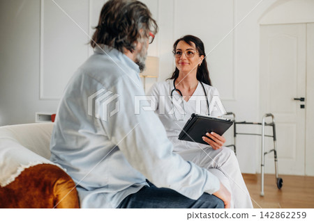 Female healthcare professional in white coat consults with male patient sitting on a couch in a well-lit room with a walker visible in the background Female healthcare professional in white coat consults with male patient sitting on a couch in a well-lit room with a walker visible in the background 142862259