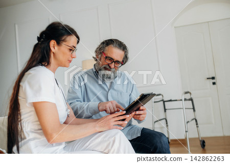 Female healthcare professional in white scrubs discusses medical information with elderly man in blue shirt, sitting in bright room with walker nearby Female healthcare professional in white scrubs discusses medical information with elderly man in blue shirt, sitting in bright room with walker nearby 142862263