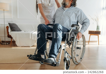 Healthcare professional assisting elderly man in wheelchair inside modern living room with comfortable furniture and natural light streaming through windows 142862268