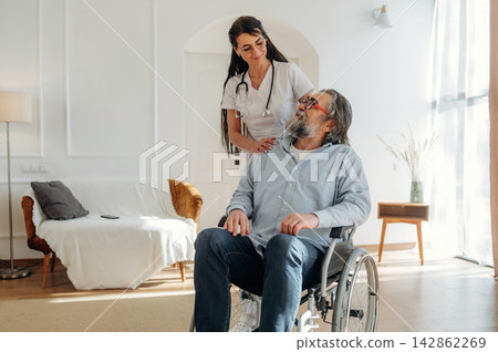 Female healthcare professional assists elderly man in wheelchair inside a bright living room with modern furniture and natural light streaming through large windows Female healthcare professional assists elderly man in wheelchair inside a bright living room with modern furniture and natural light streaming through large windows 142862269