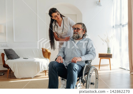 Female healthcare worker assists elderly man in wheelchair at home, bright living room with couch and natural light streaming through large windows Female healthcare worker assists elderly man in wheelchair at home, bright living room with couch and natural light streaming through large windows 142862270
