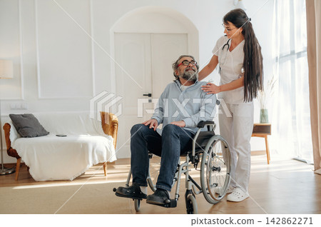Female healthcare worker in white scrubs assists elderly man in wheelchair inside a well-lit living room with modern furniture and large windows Female healthcare worker in white scrubs assists elderly man in wheelchair inside a well-lit living room with modern furniture and large windows 142862271