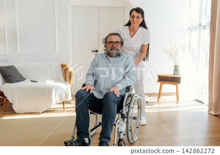 Caregiver assisting elderly man in wheelchair inside a bright, modern living room with a sofa and decorative plant in the background Caregiver assisting elderly man in wheelchair inside a bright, modern living room with a sofa and decorative plant in the background 142862272